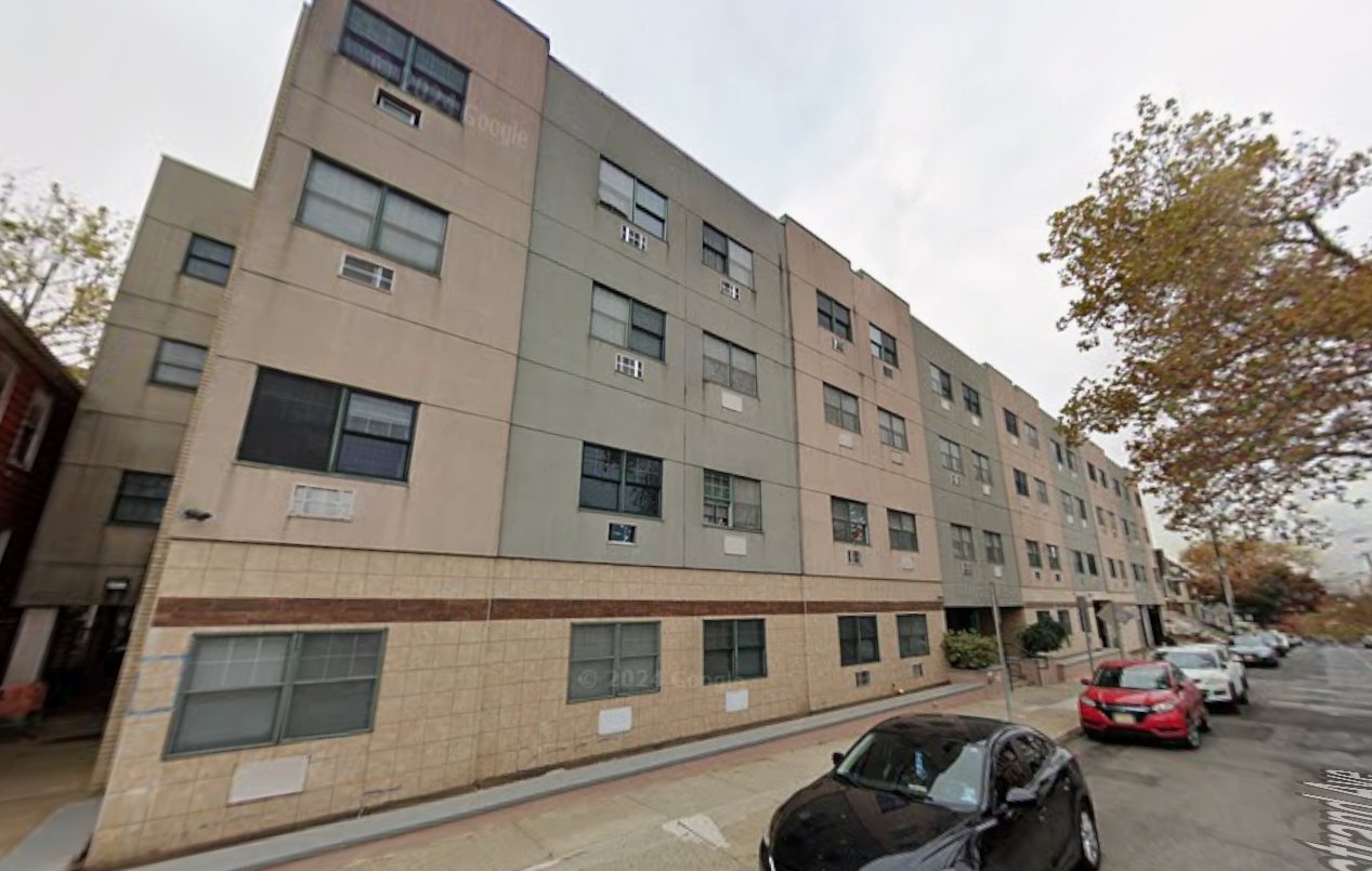 Street view of the four-story JHRC Senior Housing building on Van Nostrand Avenue, with a tan tile base, brick band, and alternating tan and gray stucco bays